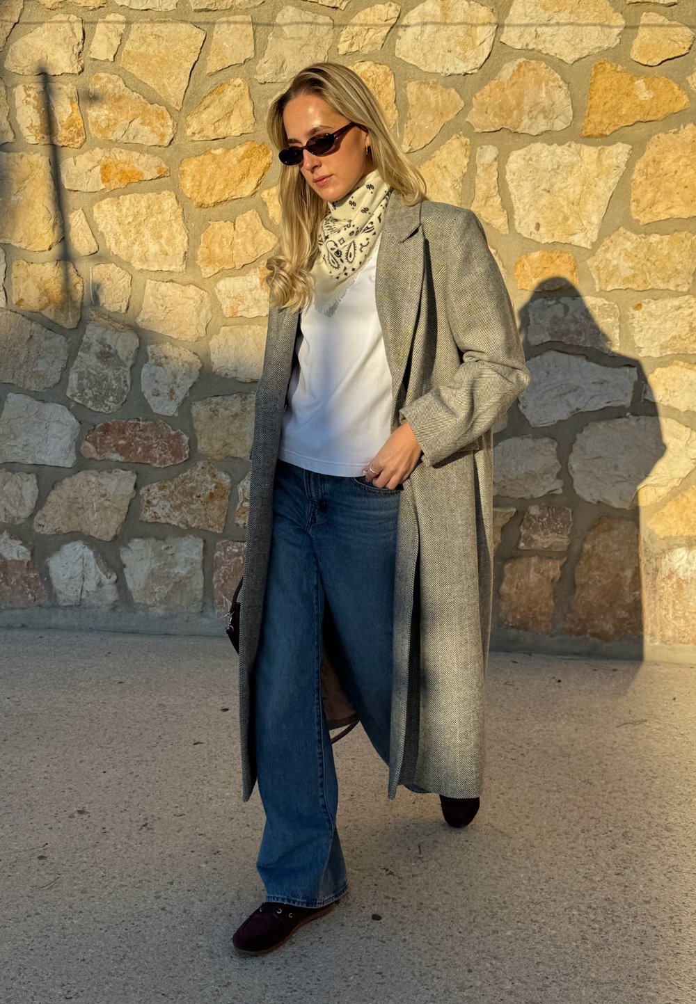 Manteau gris à chevrons porté sur une chemise blanche, associé à un jean évasé bleu moyen et des chaussures foncées. Écharpe à motifs et lunettes de soleil incluses.