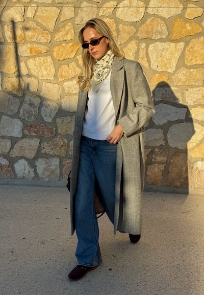 Abrigo gris de espiga llevado sobre una camisa blanca, combinado con vaqueros acampanados azul medio y calzado oscuro. Incluye bufanda con estampado y gafas de sol.