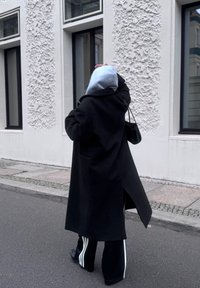 Persona con cappotto nero, cappuccio grigio e pantaloni a righe che cammina sul marciapiede accanto a un edificio bianco texturizzato con finestre con cornici nere.