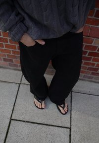 Henkilö, jolla on harmaa nyörihermosweater, mustat housut ja mustat flip-flopit, seisoo päällysteellä punatiiliseinän taustalla.