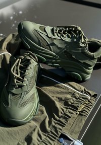 Πράσινα αθλητικά παπούτσια με επάνω μέρος από δίχτυ και καουτσούκ, χοντρή σόλα και υφές, τοποθετημένα πάνω σε ελαιοπράσινο ύφασμα με λεπτομέρειες από ελαστικ�ή ζώνη.
