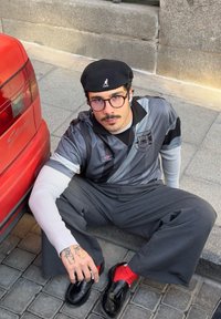 Cămașă gri de fotbal cu accente negre, pantaloni gri largi, pantofi negri și șosete roșii. Persoan ă care poartă o beretă neagră și ochelari.