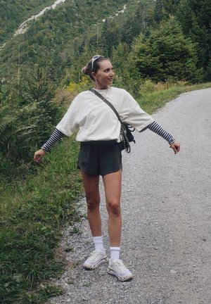 Per didelė balta medvilninė marškinė virš juodų ir baltų dryžuotų ilgųjų rankovių, derinami su žaliais šortais ir baltais sportbačiais ant žvyrkeliu.