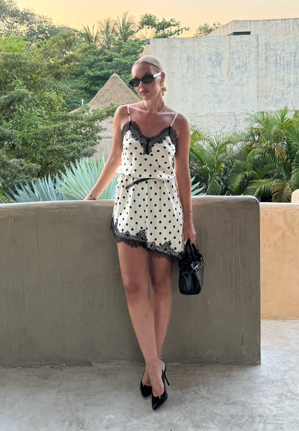 Femme portant une robe blanche à pois avec bordure en dentelle et des talons noirs, tenant un sac à main noir, debout à l'extérieur près d'un mur en béton et de la verdure.