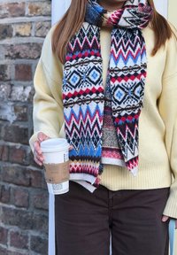 Personne portant un pull jaune, une écharpe à motifs et un pantalon foncé tient un gobelet de café à emporter près d'un mur en briques.