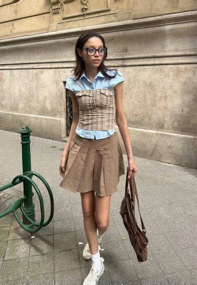 Top de corsé a cuadros con cuello de camiseta azul claro, falda plisada beige, zapatillas blancas y bolso marrón, contra un muro texturizado.