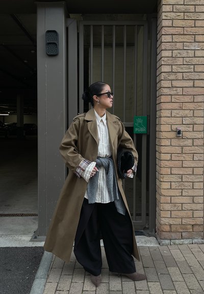 Mujer con abrigo largo marrón, camisa blanca a rayas, pantalones anchos negros, suéter gris atado a la cintura, sosteniendo un bolso negro cerca de una entrada con rejas.