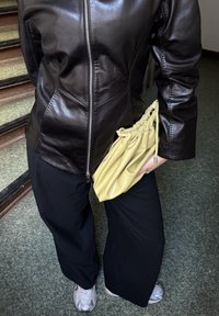 Osoba w czarnej skórzanej kurtce na zamek błyskawiczny, czarnych, szerokich spodniach, białych trampkach, trzymająca żółtą torebkę z marszczeniami przy boku w pomieszczeniu.