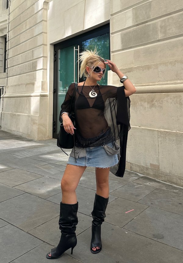 Femme portant un top noir transparent, une mini-jupe en jean et des bottes noires à talons hauts, debout sur un trottoir en ville près d'un bâtiment en pierre.