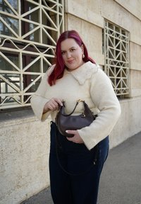 Barna bőr kézitáska ívelt formával, arany színű fém kiegészítőkkel és rövid, levehető pánttal. A modell krémszínű pulóvert visel hullámos szegélyű gallérral.