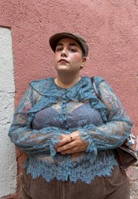 Bluză din dantelă albastră transparentă cu detalii franjurate, purtată cu pantaloni din catifea către maroniu. Ținând o geantă maro cu un inel pe mână. Fundal texturat.