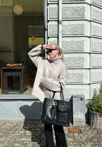 Överdimensionerad beige tröja med hög krage, svart läderhandväska med två framtaschar, stående mot en texturerad stenvägg.