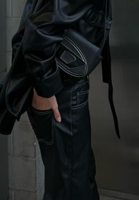 Μαύρο δερμάτινο σύνολο με γυαλιστερή επιφάνεια, oversized πουκάμισο και εφαρμοστά παντελόνια με λευκό αντίθετο ράψιμο; μια μαύρη τσάντα με γεωμετρικά διακοσμητικά στοιχεία.