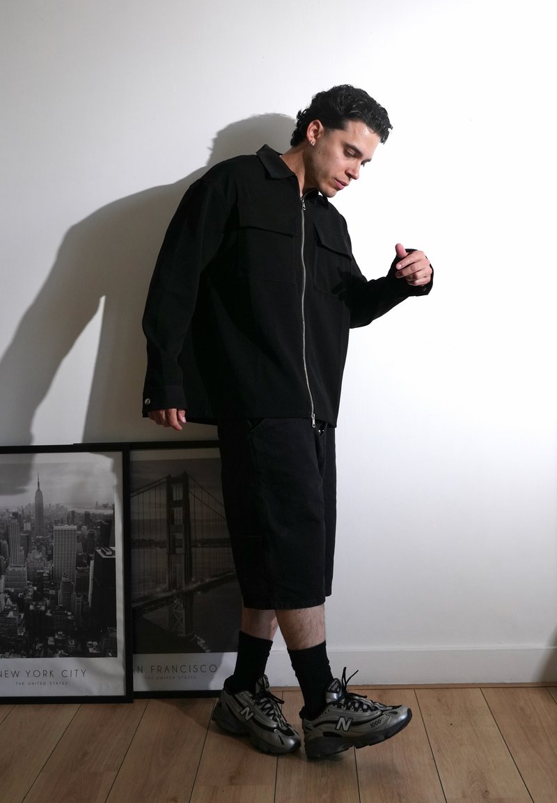 Homme en veste noire zippée, short et baskets grises, debout regardant vers le bas près de photos encadrées de paysages urbains sur un sol en bois.