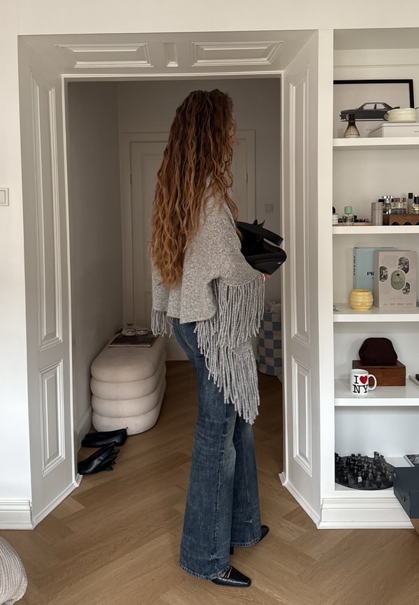 Femme aux longs cheveux bouclés portant un pull gris à franges et un jean évasé, debout dans une porte tenant un sac noir dans une pièce moderne.