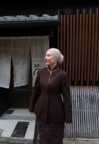 Kvinde iført beige hovedbeklædning, mørkebrun blazer og blonde nederdel står og smiler nær trælameller og en stofdør med japanske tegn.