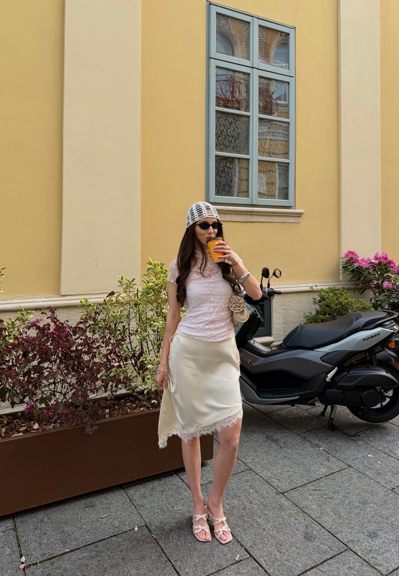 Femme en haut en dentelle blanche, jupe en soie et chapeau en filet boit une boisson orange près d'un scooter et de plantes en pot dans un cadre urbain.
