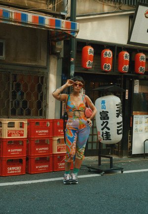 Conjunto de dos piezas colorido y con estampados que presenta gráficos vibrantes, usado con gafas de sol y zapatillas. El fondo incluye cajas rojas y faroles.
