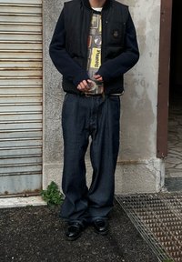 Personne portant un gilet noir par-dessus un pull à motifs, un pantalon ample rayé et des chaussures noires, debout sur une surface pavée près de l'entrée d'un bâtiment.