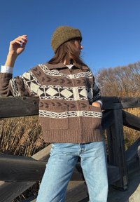 Femme portant un pull à motifs marron et un bonnet olive, appuyée contre une clôture en bois avec de l'herbe sèche et un ciel bleu clair en arrière-plan.