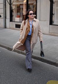 Beige trenchcoat over een bruin vest en een blauwe shirt, grijze broek, zwarte zonnebril, terwijl hij/zij een kleine zwarte handtas vasthoudt en over het trottoir wandelt.