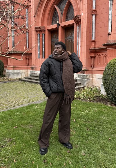 Hombre con chaqueta negra, pantalones anchos marrones y gran bufanda marrón de pie sobre el césped frente a un edificio de ladrillo rojo con puertas arqueadas de madera.