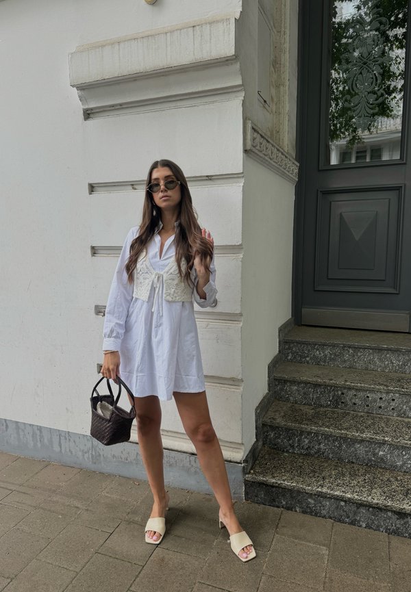 White long-sleeve shirt dress with a crochet vest, light beige heeled mules, black woven handbag, and sunglasses; standing on stone steps.