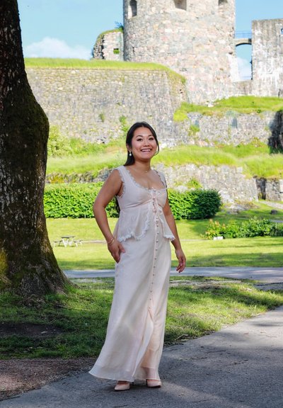 Largo vestido rosa claro con bordes de encaje y botones en la parte delantera, complementado con tacones a juego. Fondo natural con vegetación y ruinas de piedra.