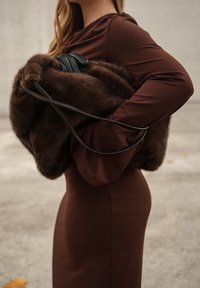 Brun pelscape holdt under armen, med mørke læderdetaljer og en glat, tætsiddende brun kjole. Tekstureret luksus med minimalistisk design.
