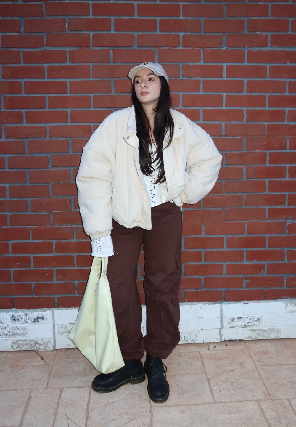 Veste matelassée oversize blanche, pantalon ample marron, haut en dentelle blanc, bottes noires et casquette beige. Porte un sac fourre-tout crème. Fond en briques.