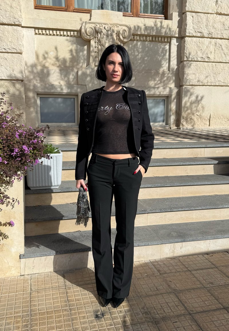 Femme aux cheveux noirs courts portant une veste courte noire et un pantalon, debout sur des marches en pierre en tenant une petite pochette argentée scintillante.