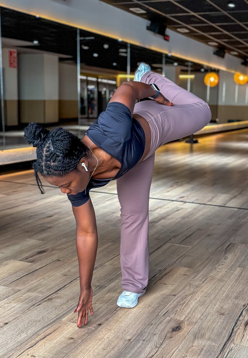 Athletic outfit featuring a black fitted top and light purple leggings, with white trainers. The person is in a balancing pose on wooden flooring.