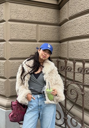 Mujer con abrigo de piel beige, gorra azul de LA, sosteniendo un batido verde, de pie frente a una pared de piedra texturizada con una barandilla de hierro.