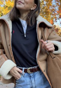 Braune Schaffelljacke mit sandfarbenen Akzenten, schwarzes Zip-Up-Oberteil mit rotem Logo, blaue Jeans und ein brauer Gürtel mit goldener Schnalle.