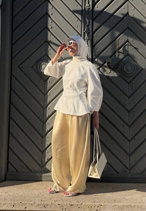 Frau mit weißem Hijab, übergroßem weißem Oberteil, cremefarbenen satten weiten Hosen, Sonnenbrille und weißen Sandalen, die eine weiße Handtasche an einer großen Holztür hält.