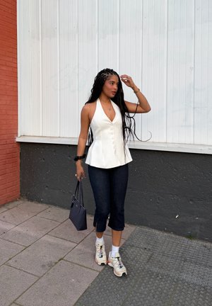 Mujer con el pelo largo trenzado, que lleva una camiseta blanca sin mangas, pantalones negros recortados, zapatillas deportivas y sostiene un bolso azul marino mientras camina al aire libre.