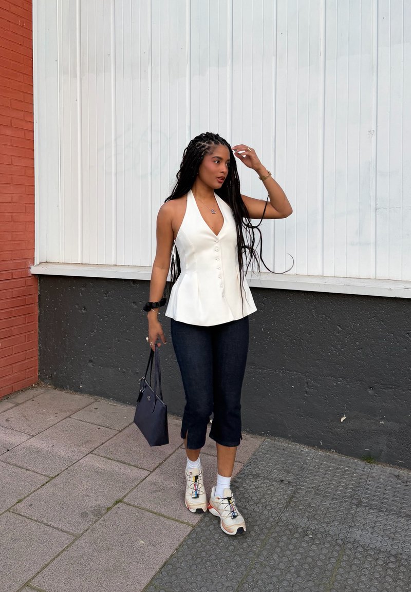 Woman with long braided hair wearing a white sleeveless top, black cropped pants, sneakers, and holding a navy handbag while walking outdoors.