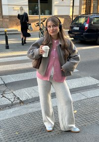Jonge vrouw met headphones drinkt koffie terwijl ze op een stadsstoep bij een zebrapad staat, gekleed in casual roze en grijze kleding.