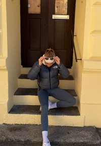 Personne portant une doudoune noire et des leggings gris assise sur des marches extérieures ajustant des lunettes de soleil blanches sur la tête.