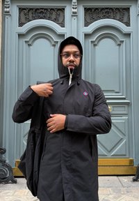 Black rain jacket with a high collar and zip-up front, featuring a logo on the chest, paired with a black hood, worn over a black shirt.