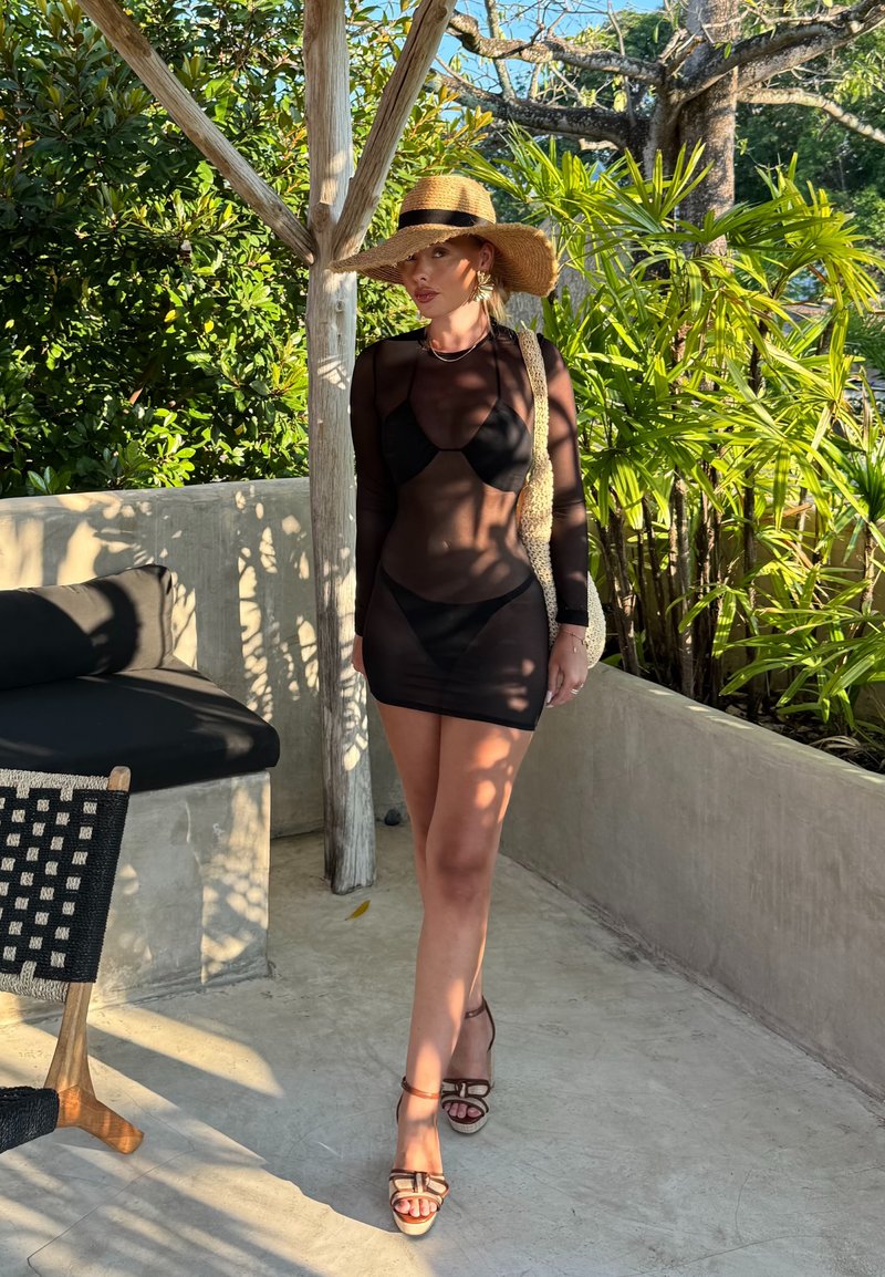 Nő fekete áttetsző ruhában, bikinivel, széles karimájú szalmakalappal, napfényben áll zöld növények és beton ülőalkalmatosságok közelében a szabadban.