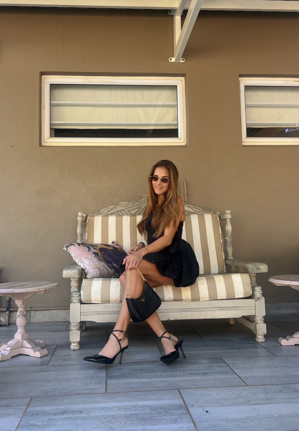 Μαύρες στιλέτο γόβες με λουράκια στο μόριο, μια μαύρη τσάντα χεριού με αλυσίδα και ένα άνετο μαύρο φόρεμα χωρίς μανίκια, είναι τοποθετημένα πάνω σε μια ριγέ καναπέ.