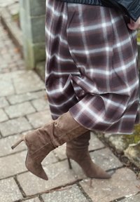 Jupe à carreaux marron avec tissu fluide, associée à des bottes à talons aiguilles en daim marron montant jusqu'aux genoux, avec bouts pointus et surfaces texturées.