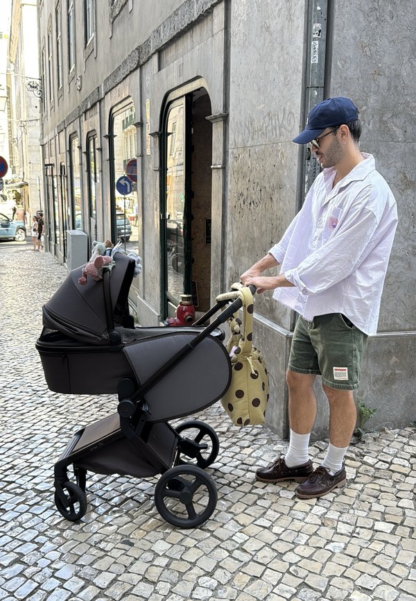 Uomo con cappellino e occhiali da sole spinge un passeggino nero su una strada lastricata accanto a un edificio in pietra con finestre e porte ad arco.