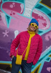 Ροζ γούνινο μπουφάν, κίτρινο πουλόβερ με λαιμόκοψη, μπλε καπέλο και κίτρινες γυαλιά ηλίου. Περιλαμβάνει μια υφασμάτινη κίτρινη τσάντα με λουράκι. Το φόντο έχει πολύχρωμο γκράφιτι.