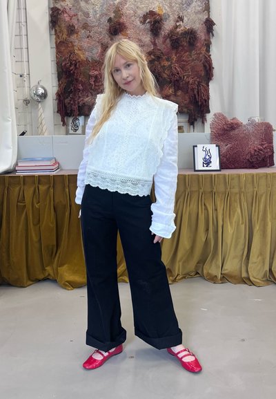 Blusa de encaje blanco con bordes rizados, combinada con pantalones de pierna ancha negros y zapatos planos rojos. El fondo texturizado presenta arte mural abstracto.