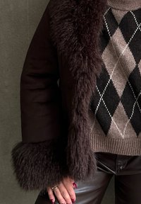 Veste extérieure marron avec un col en fourrure, portée sur un pull en motif argyle beige et noir. Elle se complète avec un pantalon en cuir noir et des ongles bien manucurés.