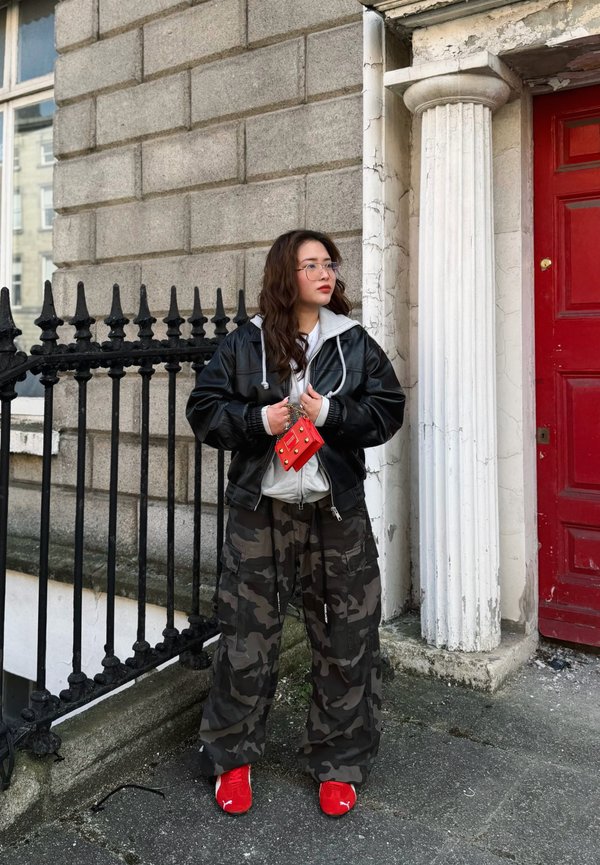 Jonge vrouw in camouflagebroek, zwarte leren jas, rode sneakers, met een klein rood tasje, staand bij een stenen gebouw met een rode deur.