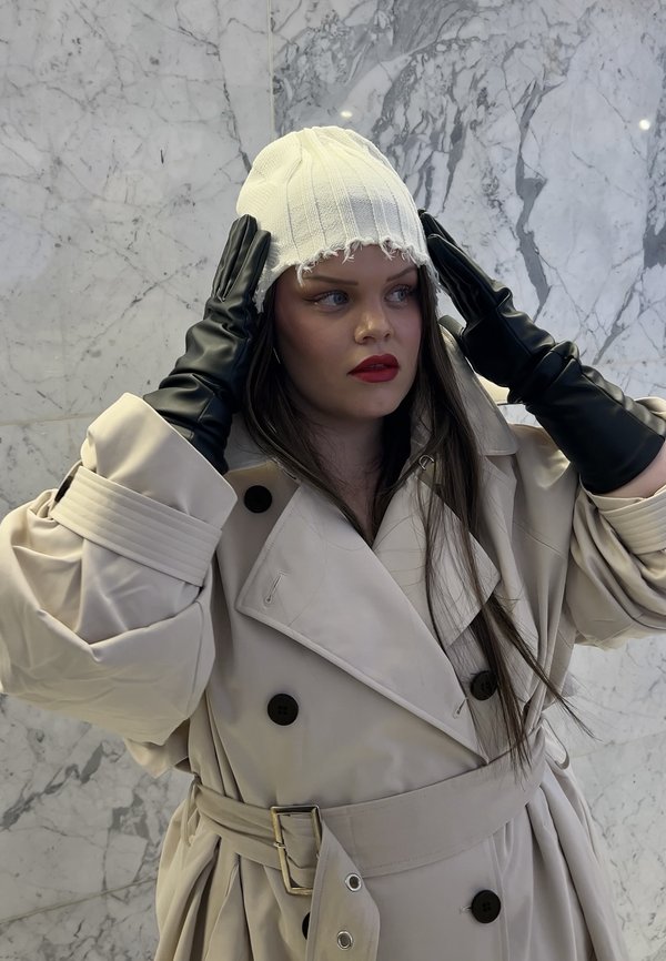 Femme en trench-coat beige et gants noirs ajustant un bonnet en maille blanc devant un mur en marbre.