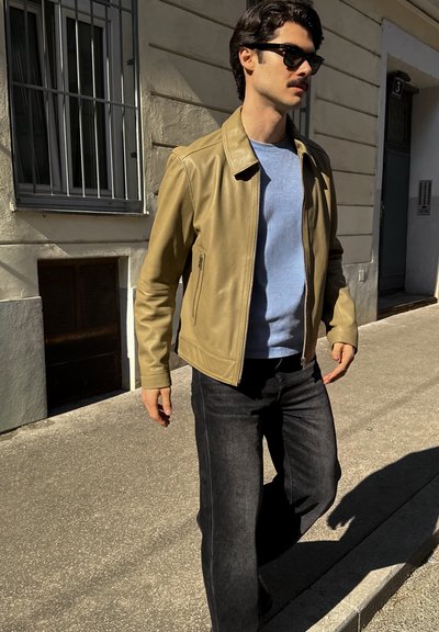 Hombre con gafas de sol, chaqueta de cuero beige, camisa azul clara y vaqueros negros caminando por una acera urbana iluminada por el sol cerca de un edificio.